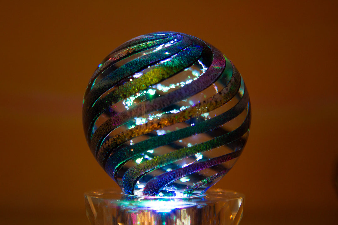 Dark dichroic rainbow glass orb with cremation ash spirals on a clear pedestal.