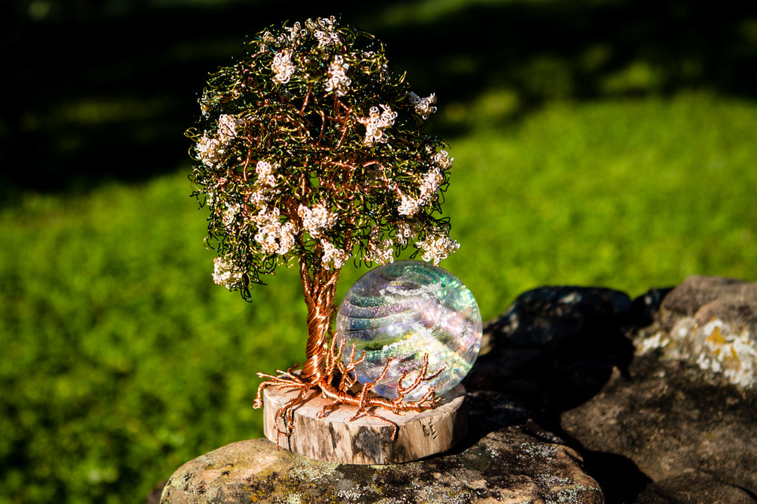 Handcrafted copper wire tree of life with white blossoms on a wooden base beside a swirled glass orb