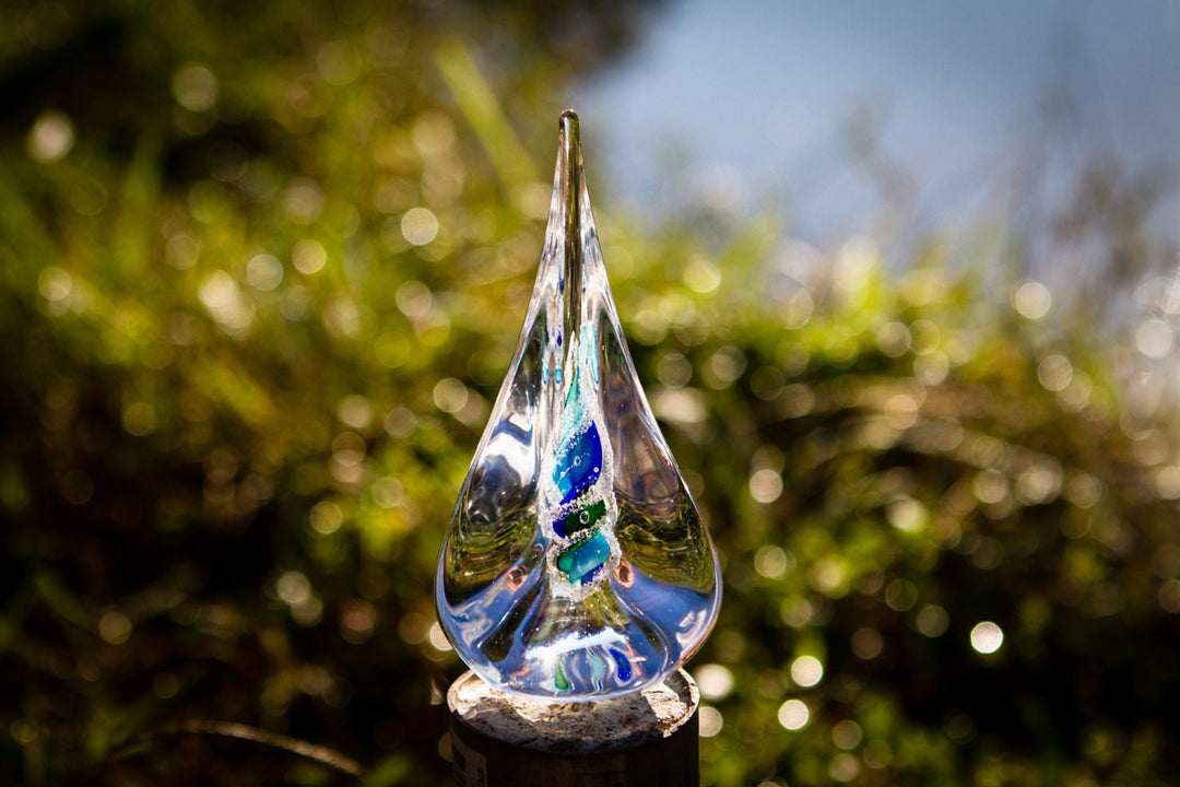 Clear teardrop pyramid with a blue green white spiral containing cremation ash shown outdoors on a rock