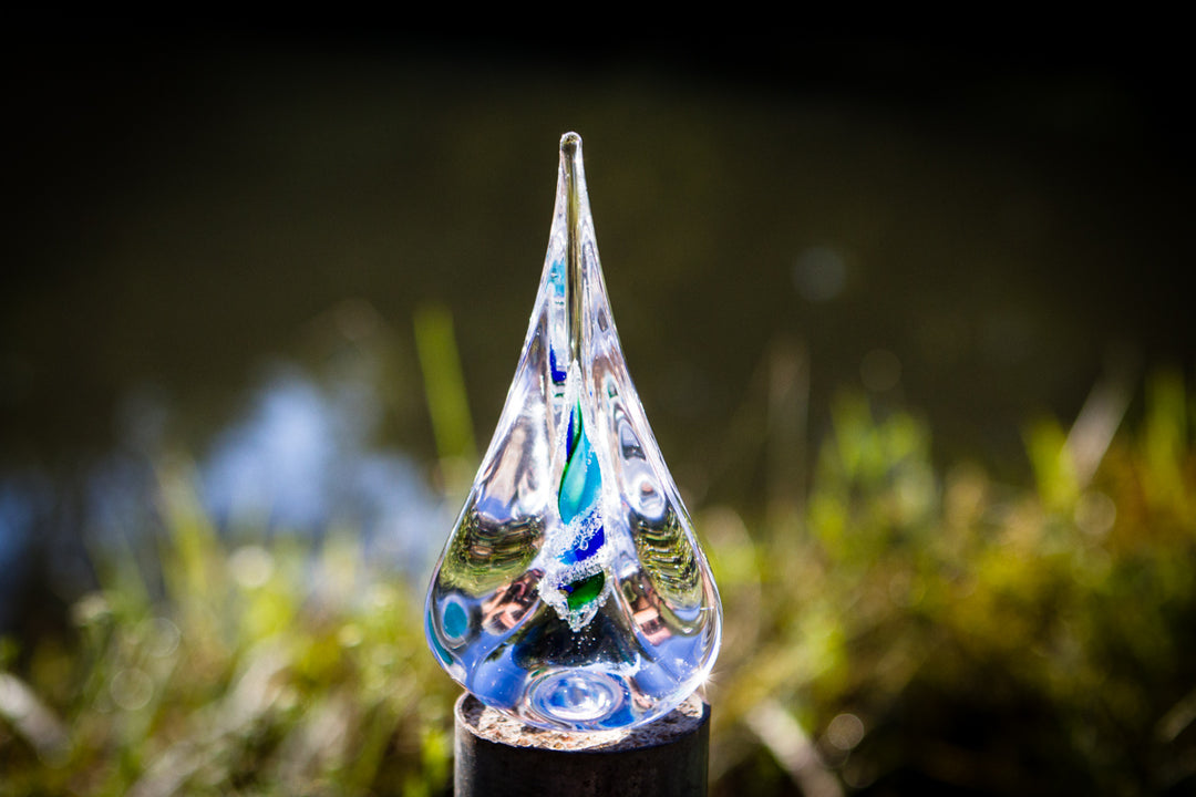 Clear glass memorial flame with spiraling blue and green ash visible inside, displayed outdoors on a dark stand.