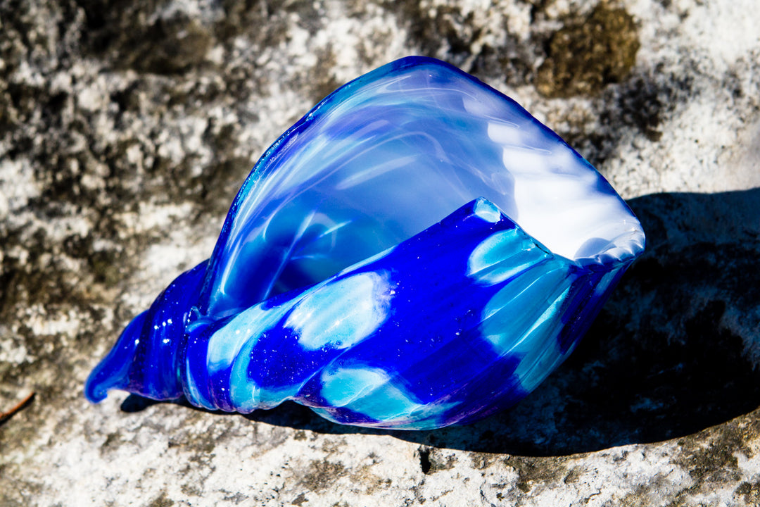 Blue glass seashell containing cremation ash resting on a rocky surface.