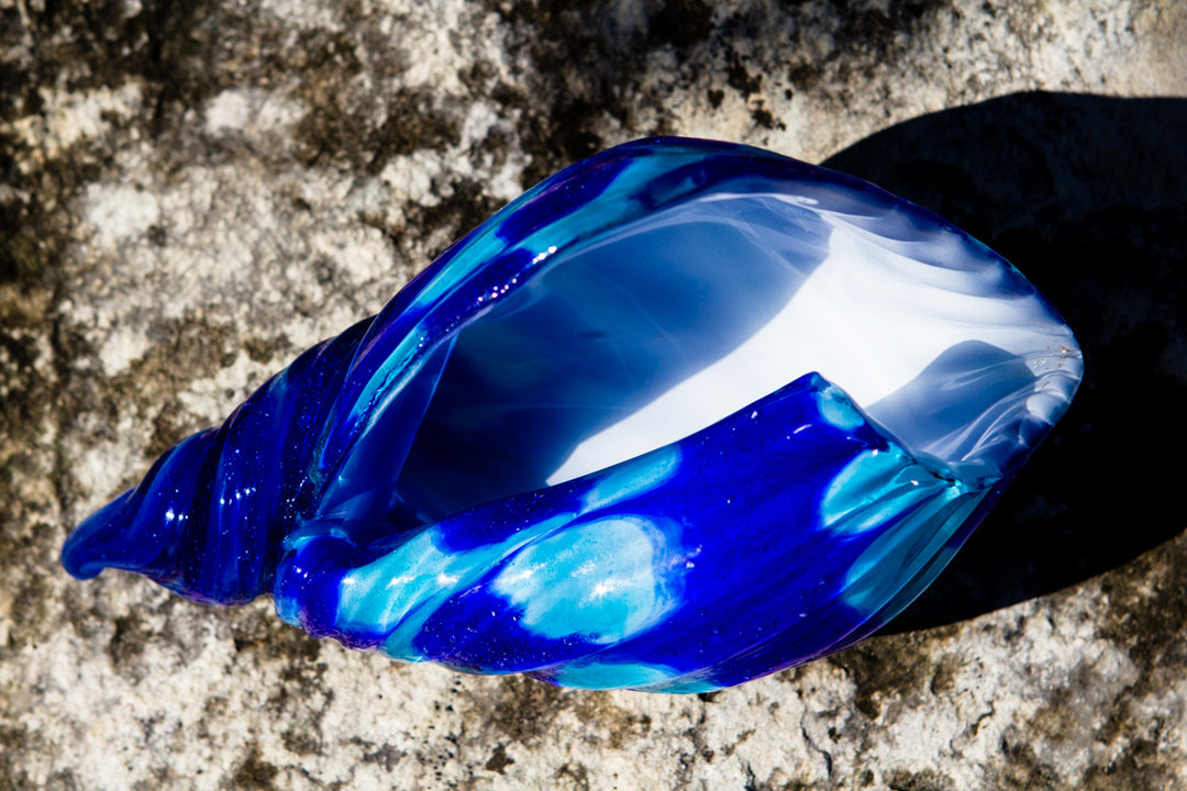 Blue glass seashell sculpture with white interior on a rocky surface