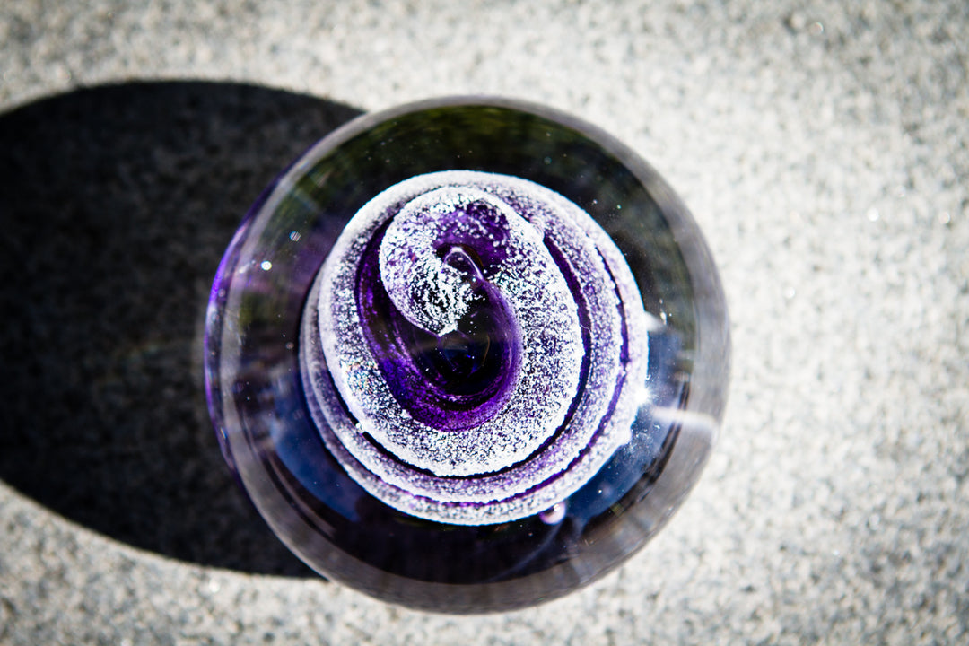 Swirl glass paperweight with cremation ash featuring purple spirals encased in clear glass on a textured surface.