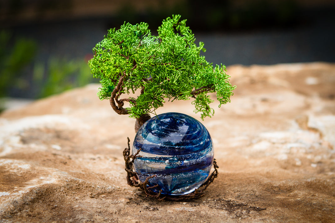 Green wire Ponderosa Tree of Life sculpture perched on a blue glass orb on a rocky surface.