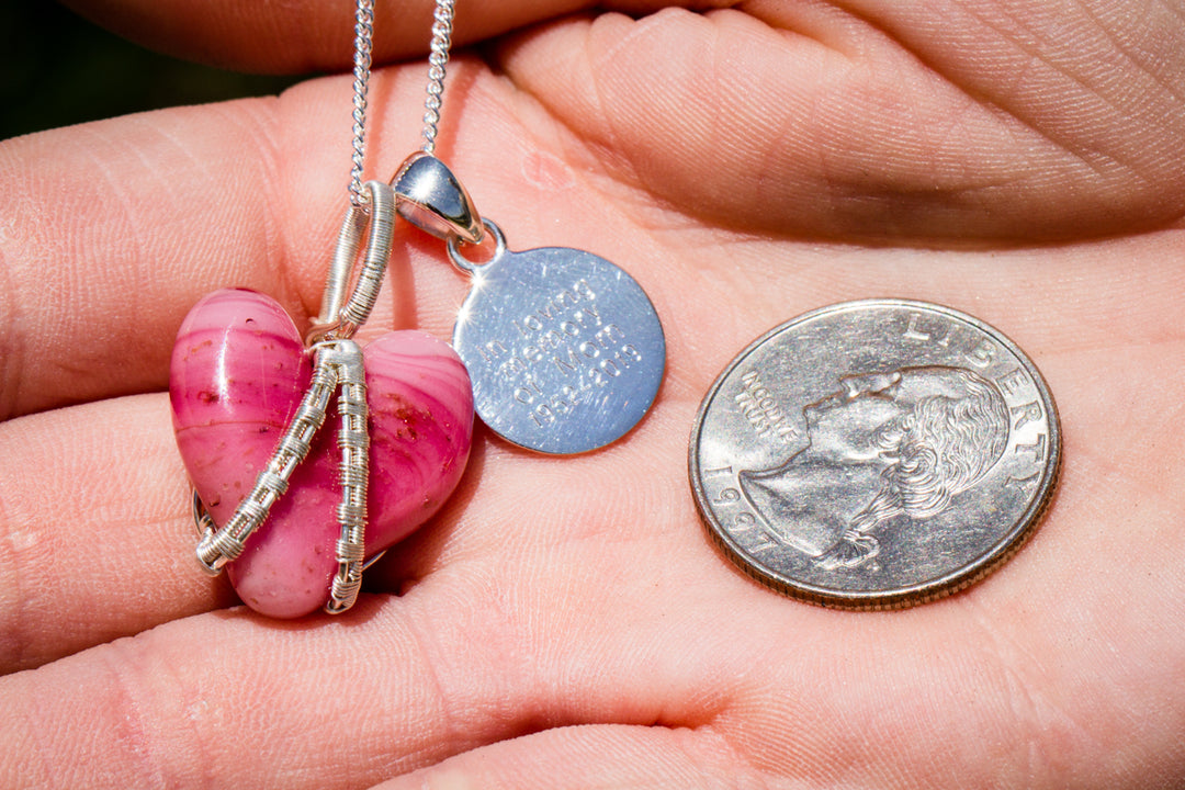 Pink glass heart pendant wrapped in silver wire on a silver chain beside a round blue charm and a quarter for scale