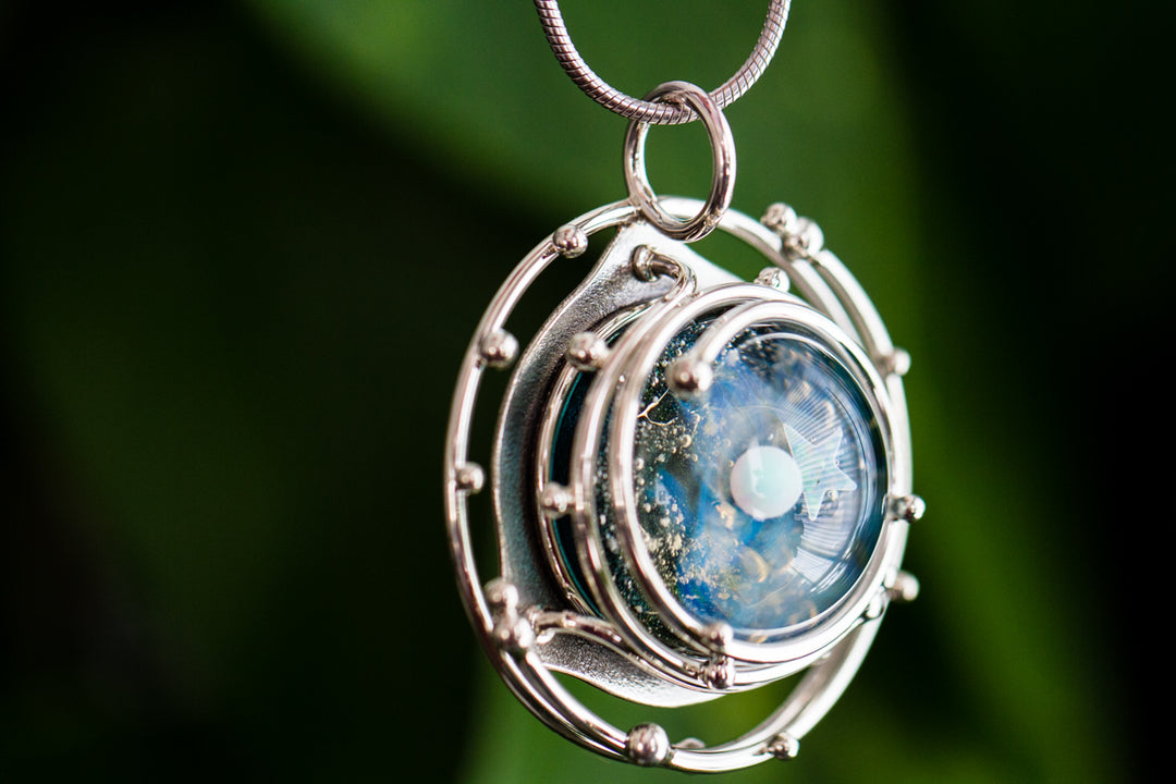 Close-up of a silver circular cage pendant with layered rings encasing a blue galaxy glass cabochon on a chain
