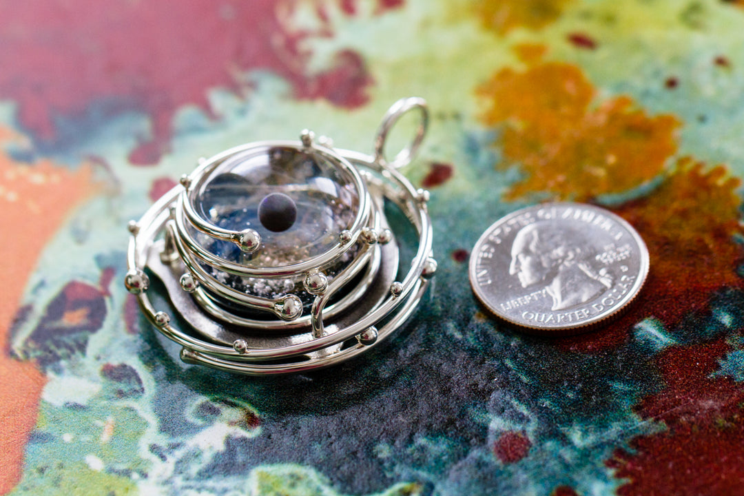 Intricate multi hoop silver pendant encasing a glass galaxy orb shown with a quarter for scale