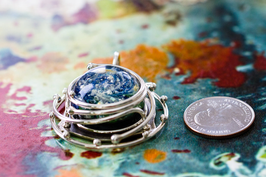 Caged glass ocean pendant with a blue and green swirling cabochon containing ashes inside a silver decorative cage, shown with a coin for scale.
