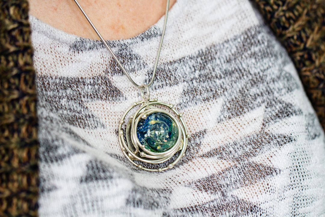 Person wears a silver spiral cage pendant housing a blue green ocean swirled glass cabochon with ashes.
