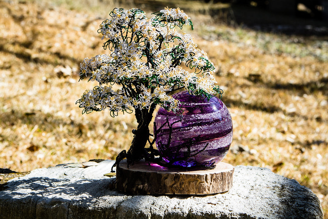 White Dogwood Tree of Life sculpture with a purple marbled orb on a wooden base outdoors