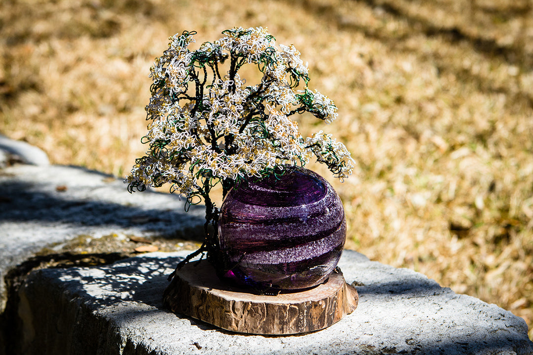 White Dogwood Tree of Life sculpture with a purple marbled orb beside it on a wooden base outdoors