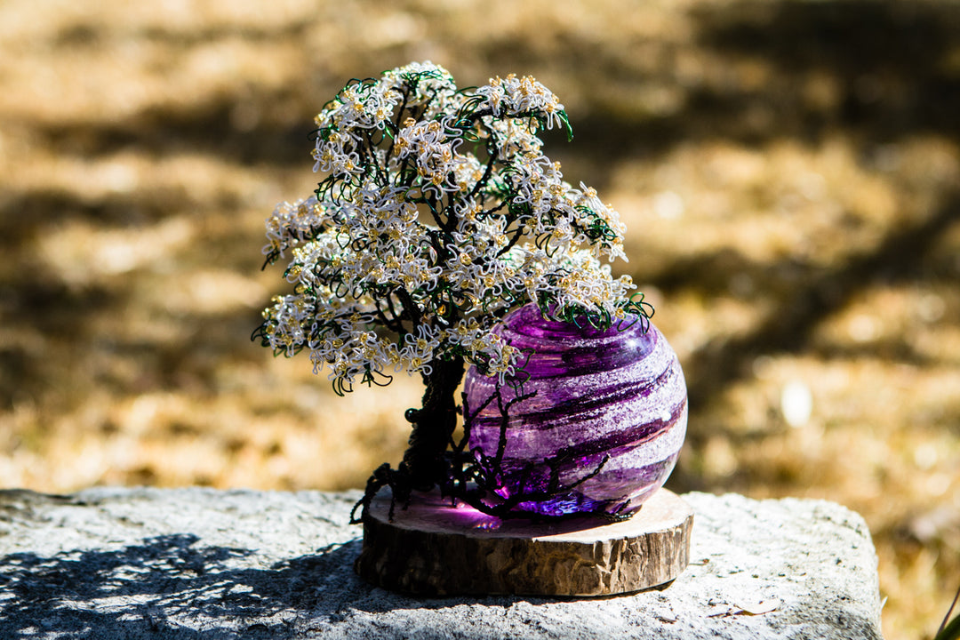 White Dogwood Tree of Life sculpture with purple marbled orb on wooden base outdoors on a stone surface.