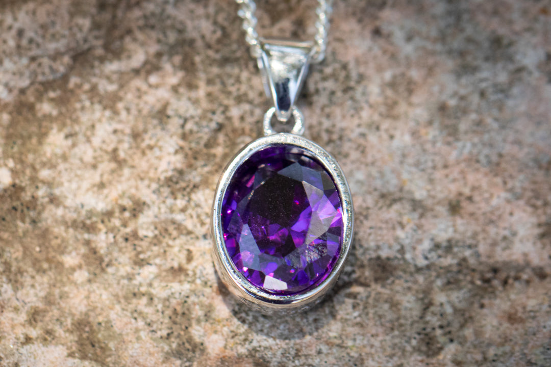 Purple oval crystal memorial pendant in a silver setting with chain on a textured rock surface