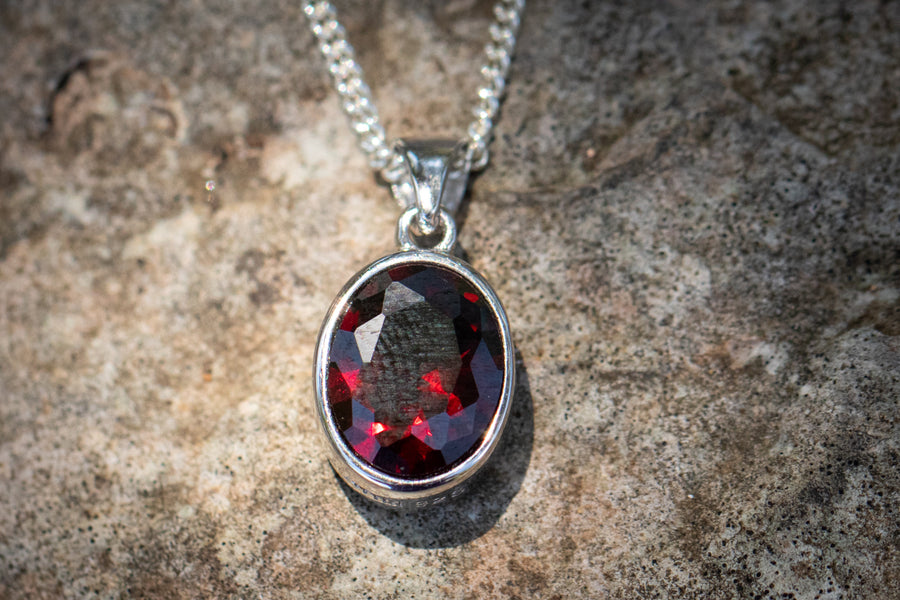 Oval red crystal memorial pendant in a silver setting with chain on a rocky surface