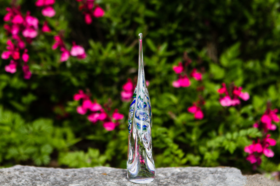 Tall teardrop glass flame drop with cremains and blue green swirl on a stone surface outdoors.
