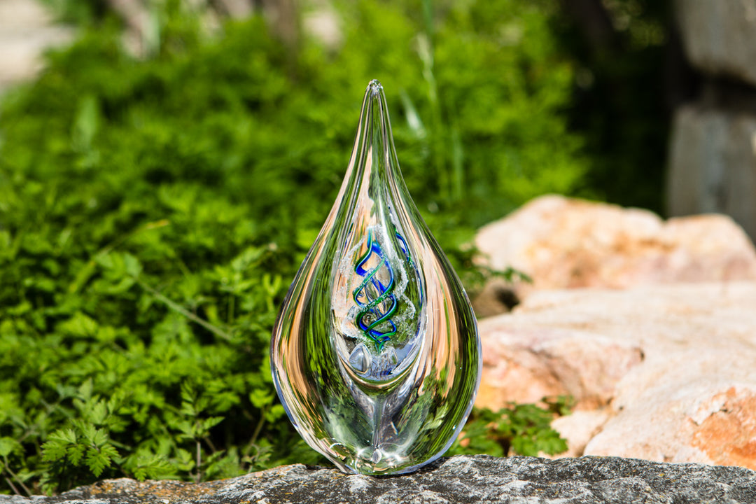 Teardrop shaped glass memorial flame drop with a vibrant inner color swirl, set on a stone surface among green plants.