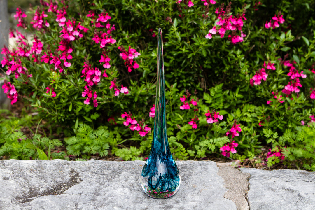 Illuminated glass angel drop infused with cremation ash outdoors on a stone surface with pink flowers in the background.