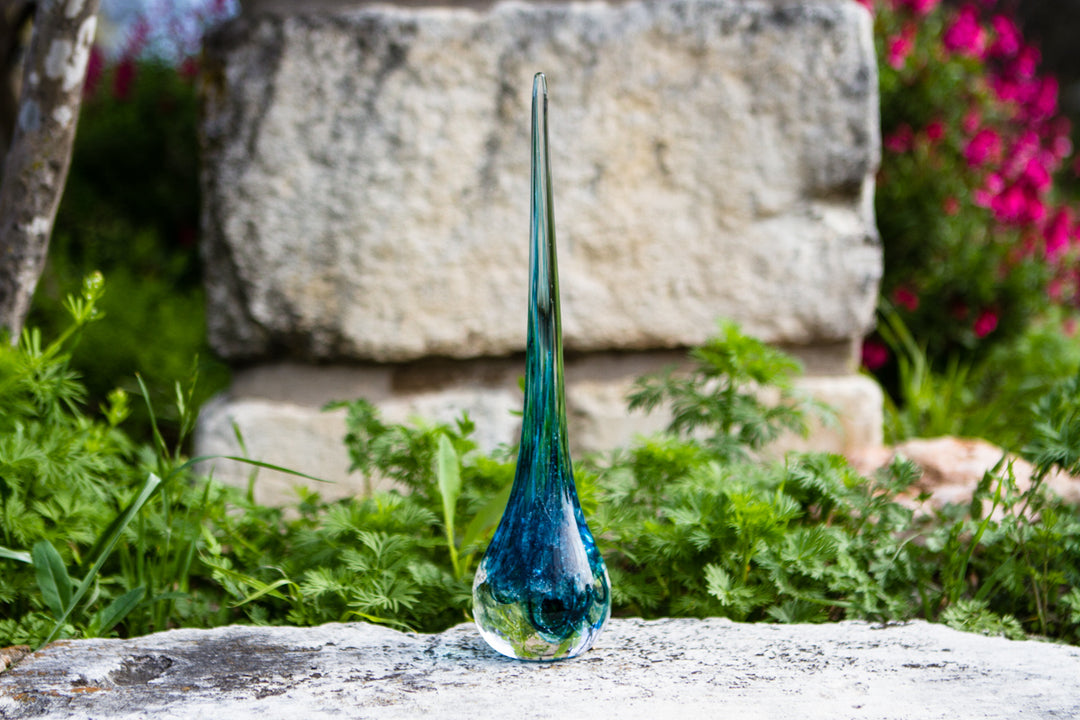 Tall blue and teal glass angel drop on a sunlit stone surface.