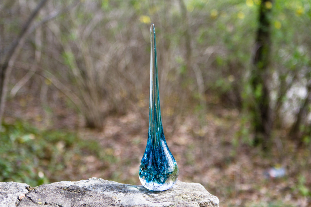 Slender blue teal glass angel drop with a curved neck outdoors on a rocky surface.