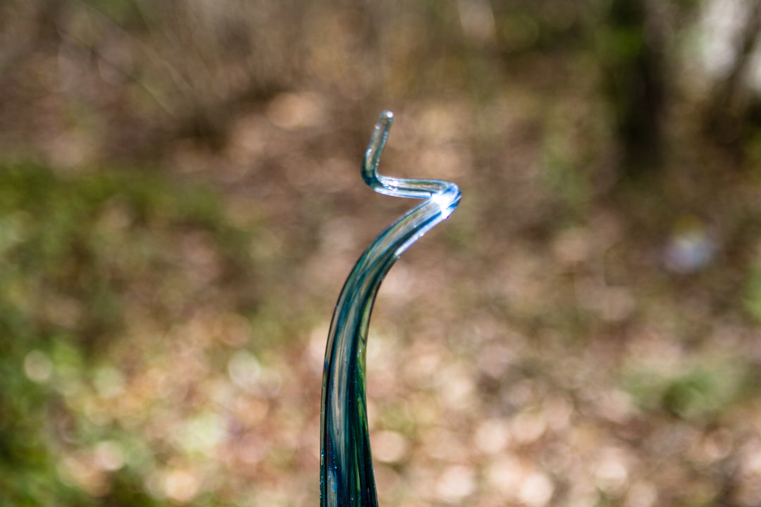 Curly top teal glass angel drop with a wavy neck resting on a stone ledge outdoors.