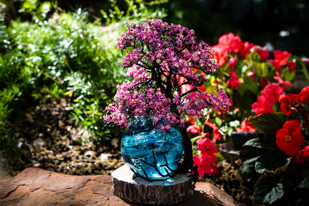 Handmade pink wire dogwood tree on a blue glass orb base mounted on a wood slice outdoors.