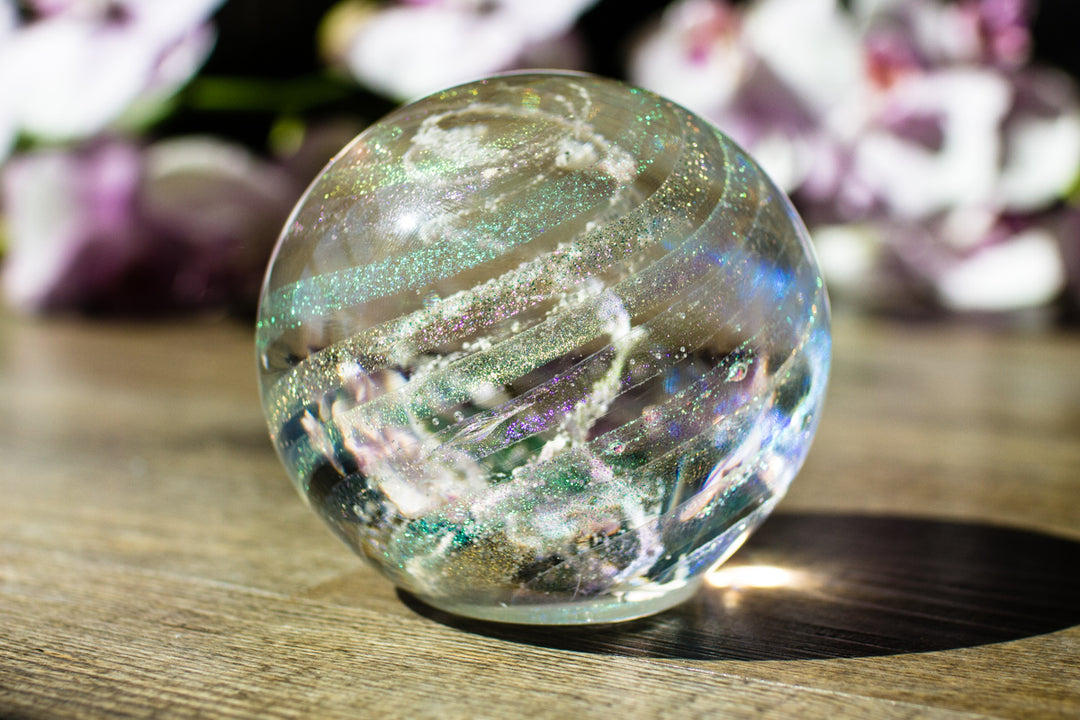 Dichroic rainbow glass orb containing cremation ashes with glittery ribbons, resting on a wooden surface.