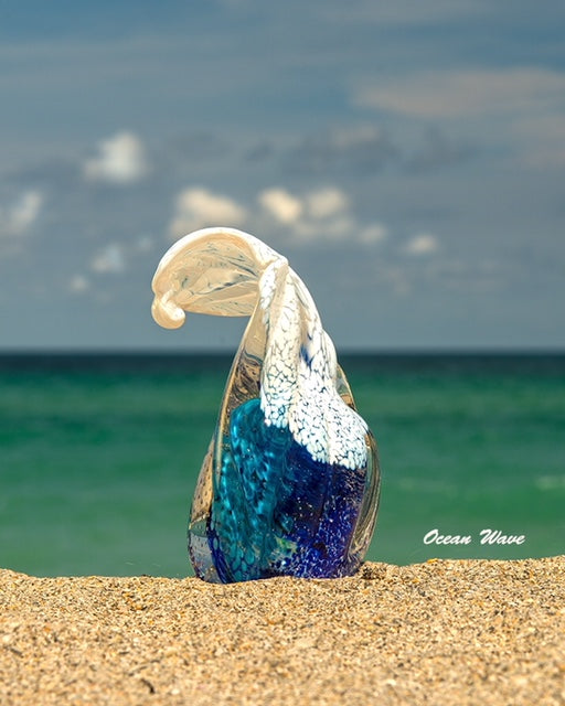 Glass ocean wave paperweight with white crest on a sandy beach with the ocean in the distance