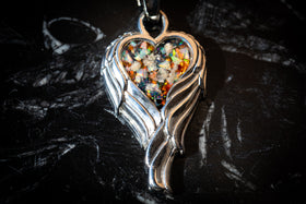 The photo depicts the angel wing heart pendant sitting on a dark background. The pendant contains orange, black, and white crushed opal along with cremains. Silver necklace for cremation ash, silver remembrance jewelry, necklace for ash