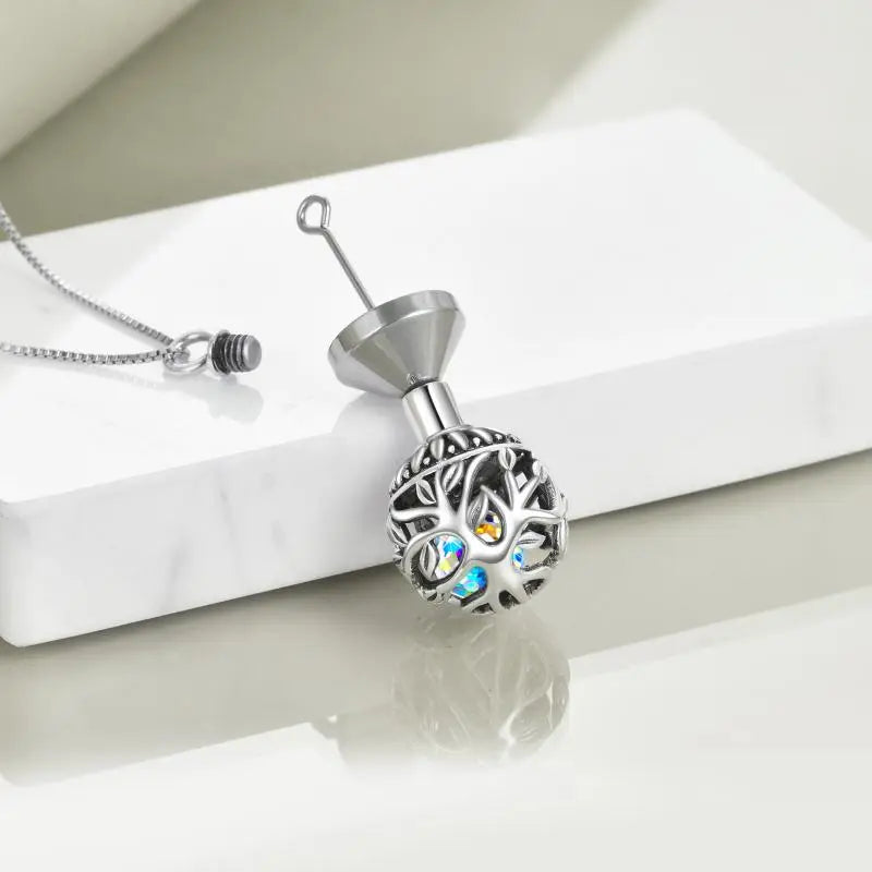 Silver ball cremation pendant with a tree of life motif and iridescent crystals inside resting on a white surface