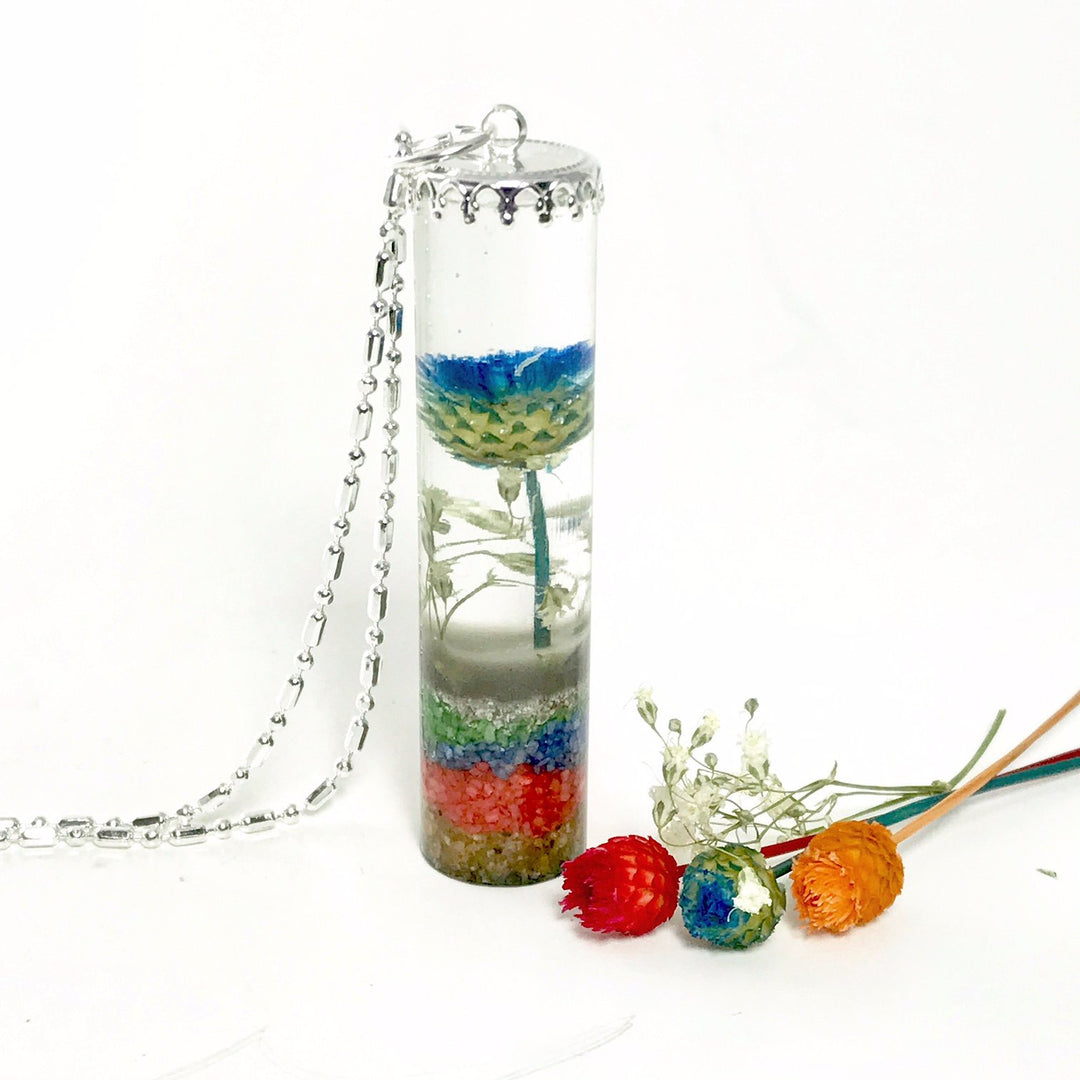 Tall cylindrical clear resin pendant housing a blue green beach flower and multicolored sand layers, shown with a silver chain