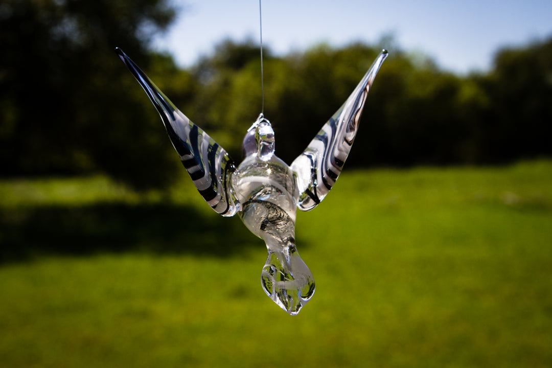 Hanging glass hummingbird viewed from below with striped wings spread over a grassy background.