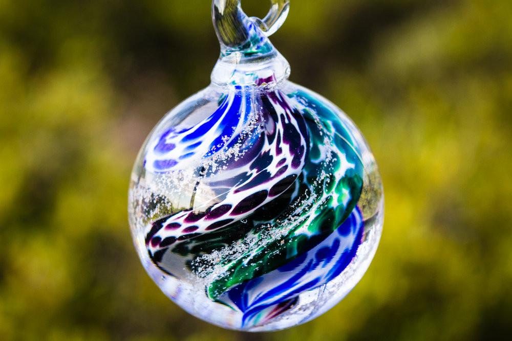 Close up of a multicolored swirled glass cremation orb in blue green and purple tones, hanging against a blurred garden background.