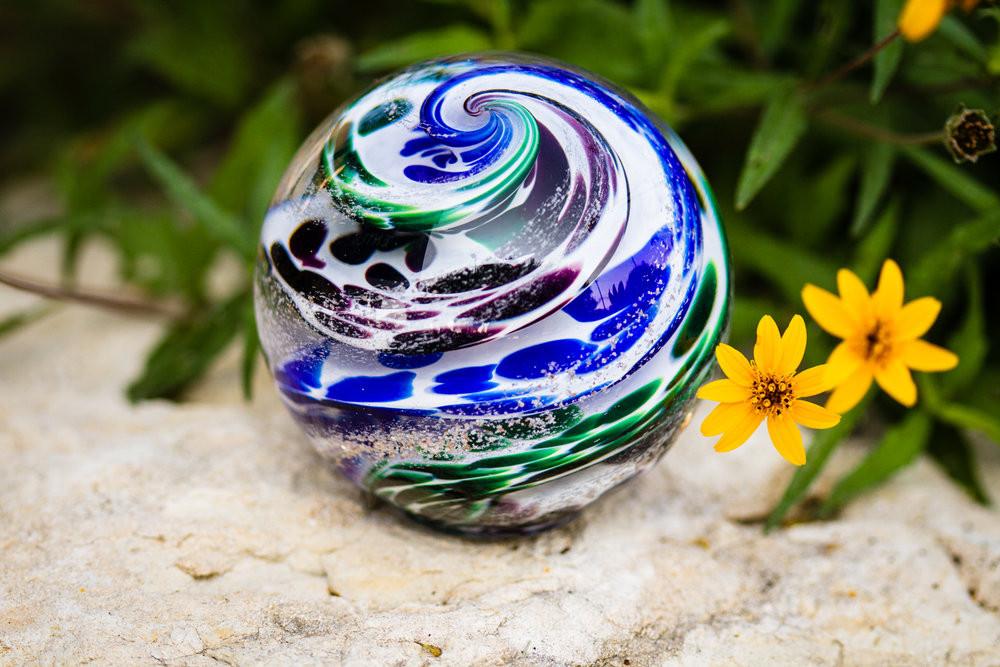 Swirled glass cremation orb with blue green white swirls sits on a stone beside bright yellow flowers.