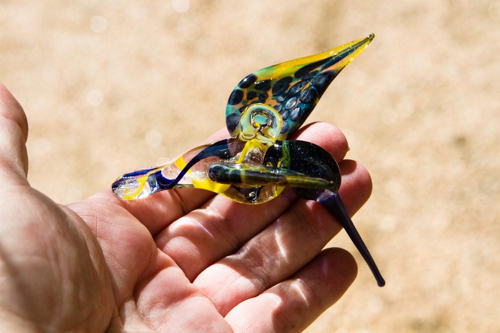 Blue and yellow cremation ashes swirl through a glass hummingbird with a hanging hook.