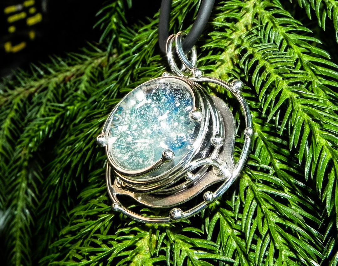 Caged glass galaxy pendant containing a blue green cabochon on a black cord.