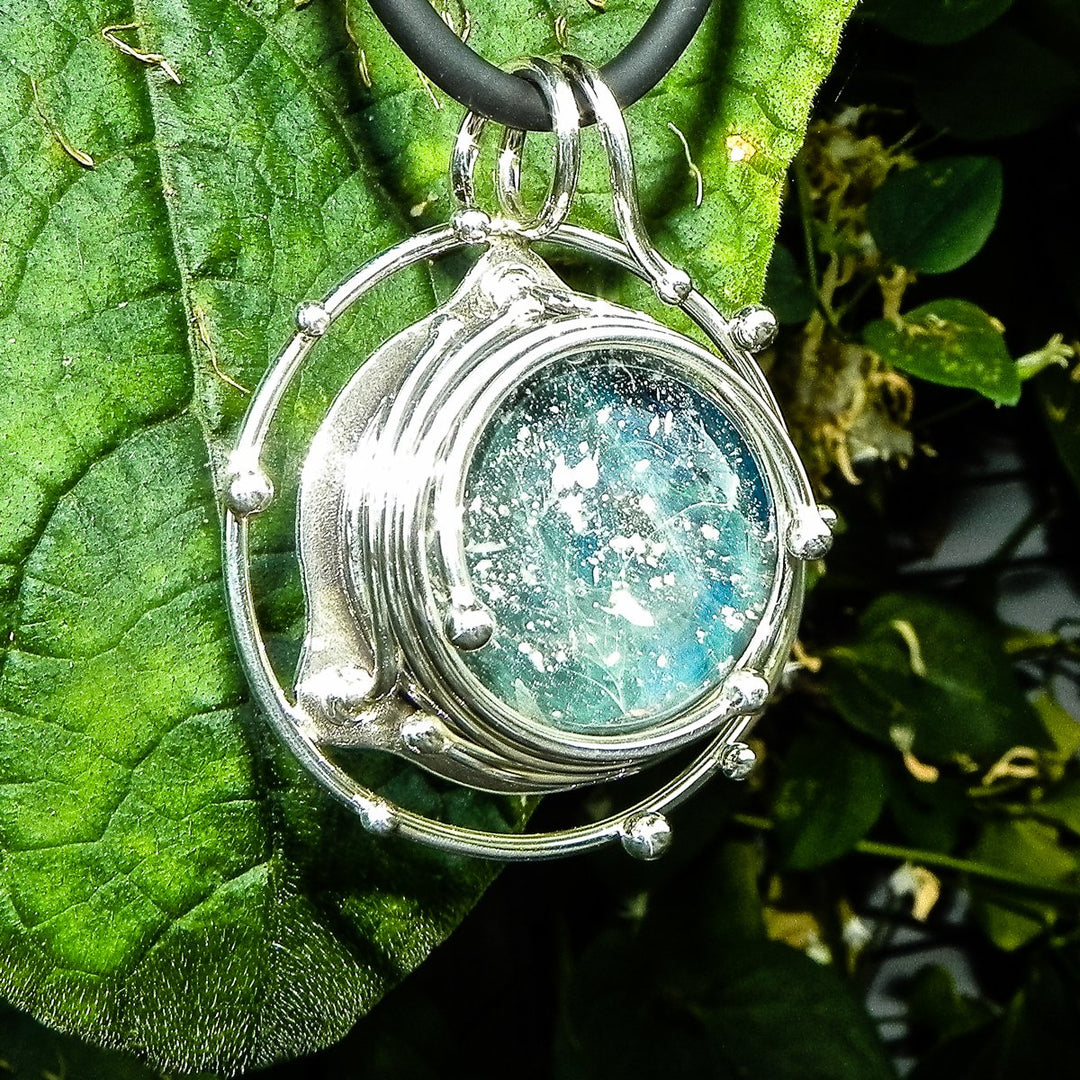 Close up of the caged glass galaxy pendant with a blue green orb in a silver frame against green foliage.