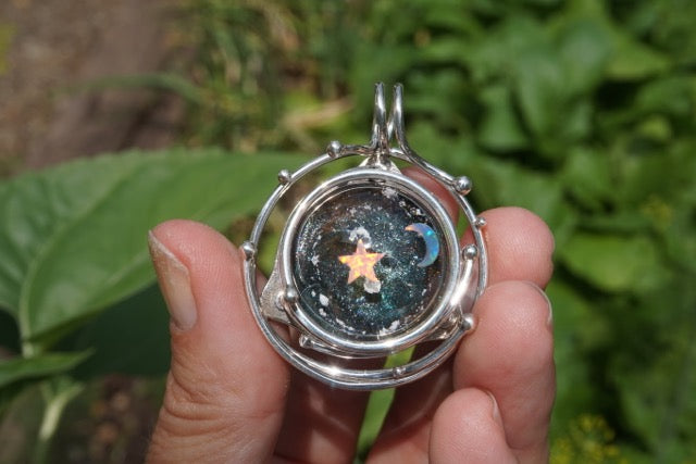 Hand holding a silver galaxy pendant with a glass cabochon displaying a star and crescent moon in a starry space scene