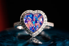 Bedazzled Heart Cremation Ring with blue and pink selection of crushed opal. The ring is sitting atop a glass surface, with the stone facing the viewer. Sterling Silver Ring for Ash, Sterling Silver Memorial Jewelry, Remembrance Jewelry, Silver Ring