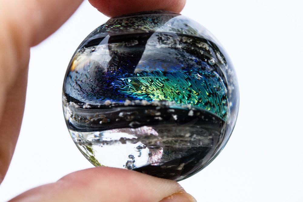 Dichroic glass twist marble infused with cremation ashes held between fingers showing iridescent blue green swirls.