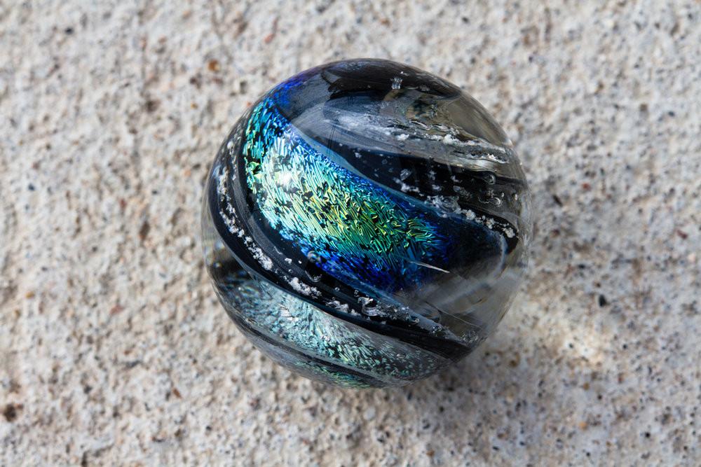 Round dichroic glass cremains marble with black edge and rainbow and blue gold copper swirls encasing ashes that will roll.