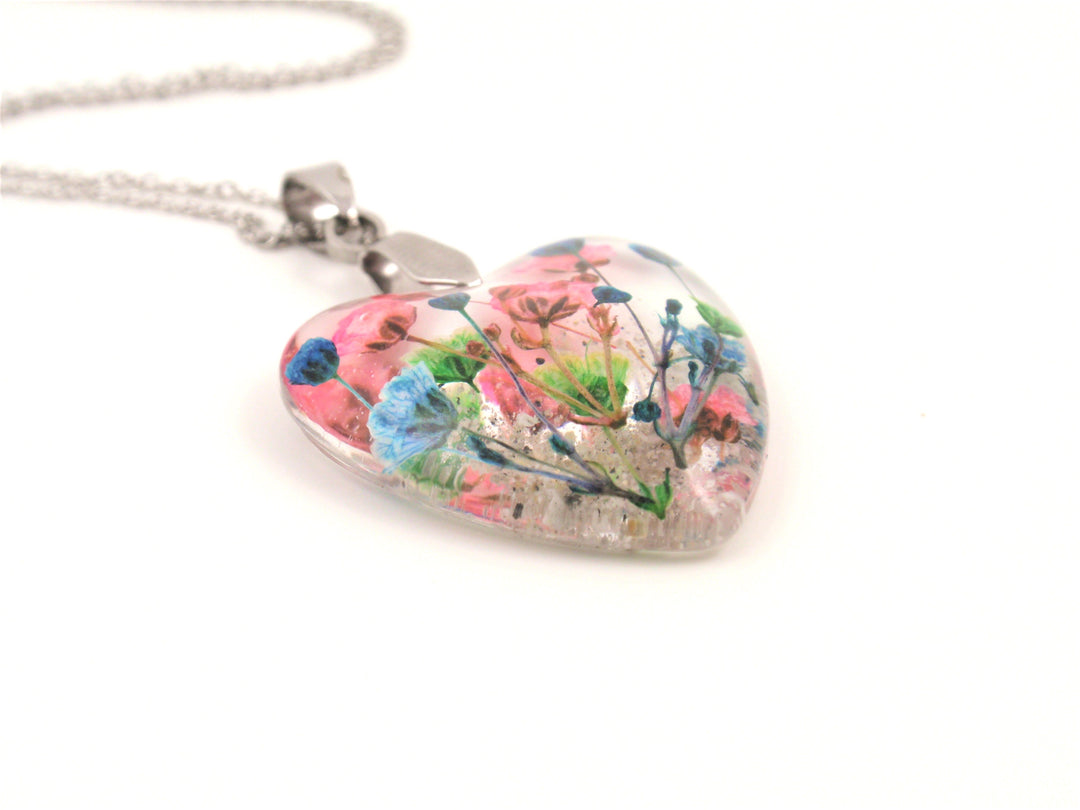 Close up of heart shaped resin pendant showing pink and blue dried flowers on a silver chain