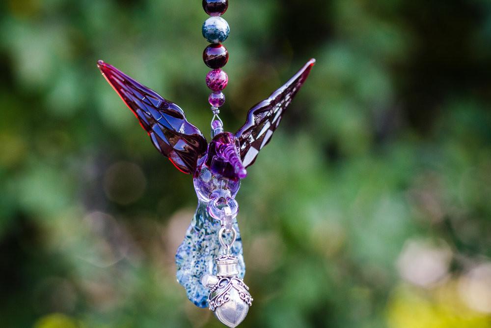 Purple maroon glass hummingbird pendant with beaded strand and sterling silver keepsake urn against a garden background