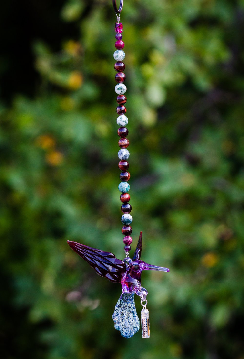 Beaded hanging charm with purple hummingbird pendant and small sterling silver keepsake urn against a garden background