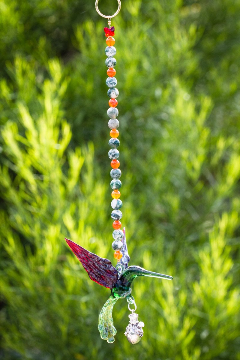 Close-up of the hummingbird pendant with a beaded strand and a decorative silver keepsake urn against a green background