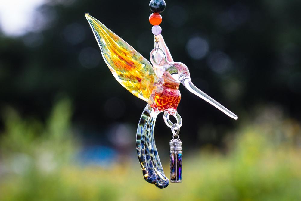 Close up of a glass hummingbird pendant with a yellow orange wing and blue speckled tail suspended beside a string of multicolor beads