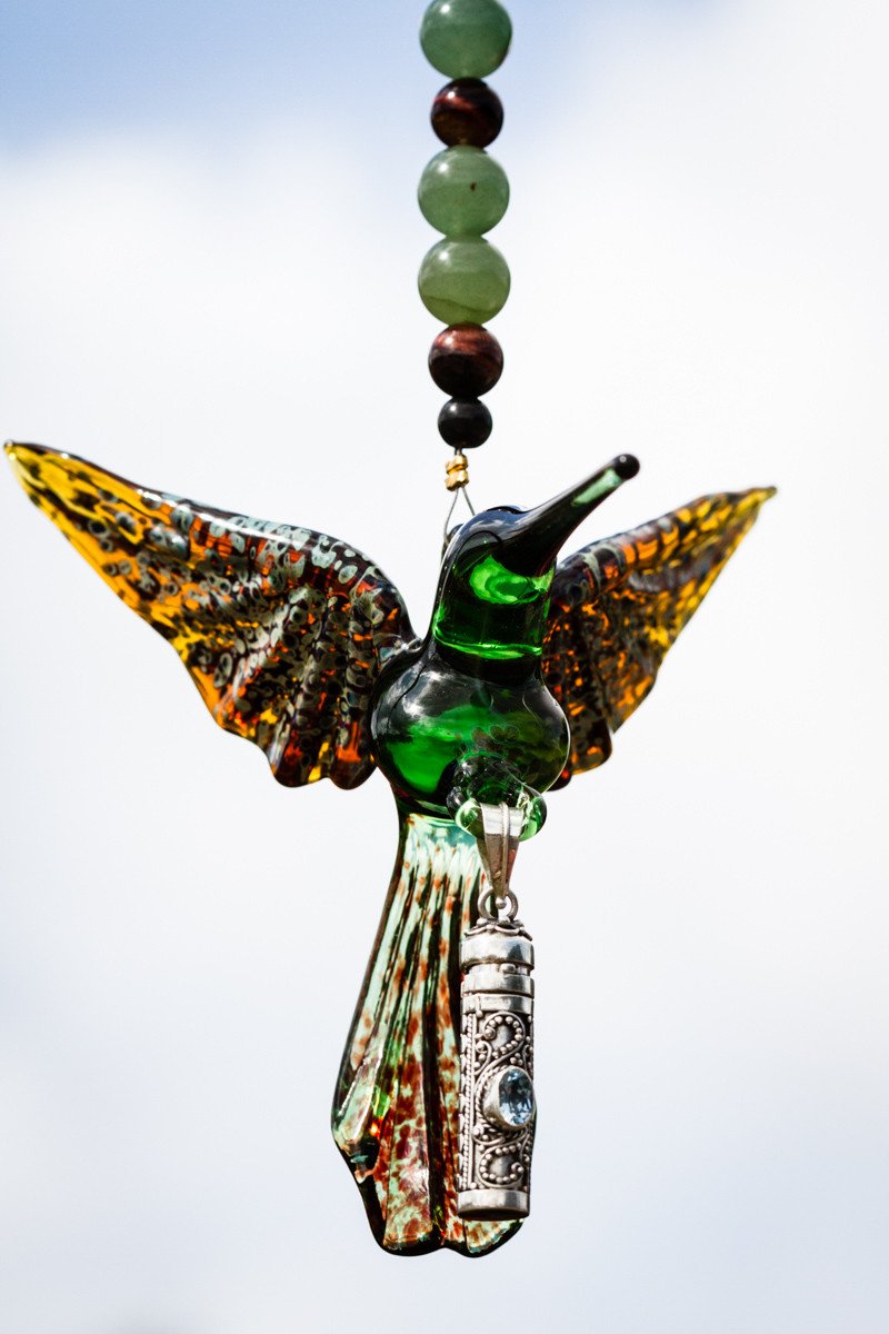 Green hummingbird pendant with spread wings and an ornate silver keepsake urn hanging below against a clear blue sky.