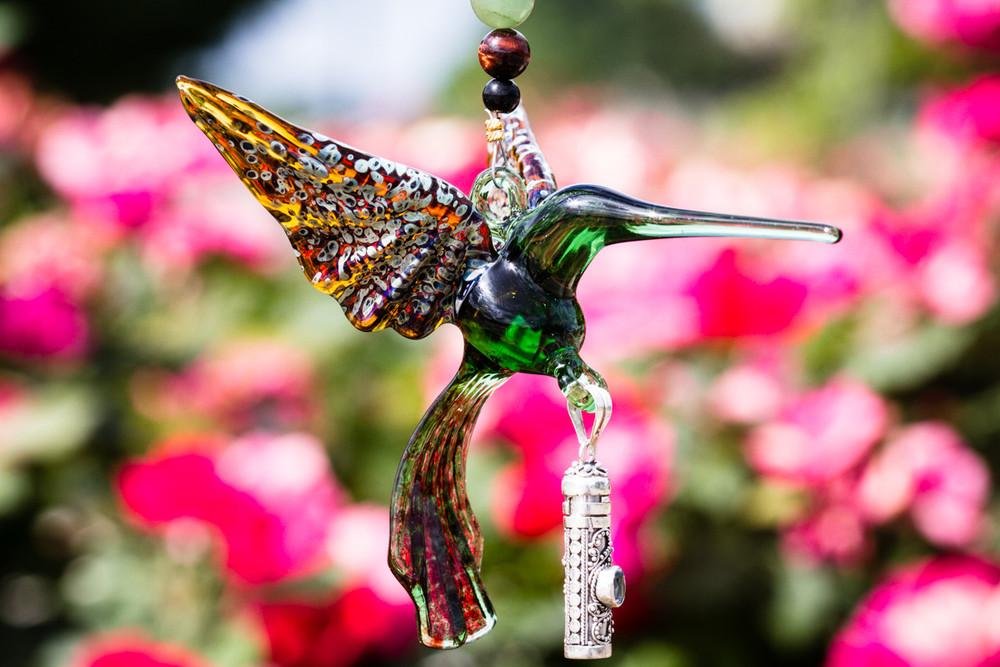 Green glass hummingbird with a silver keepsake urn pendant and red tiger eye and aventurine beads against a colorful garden backdrop.