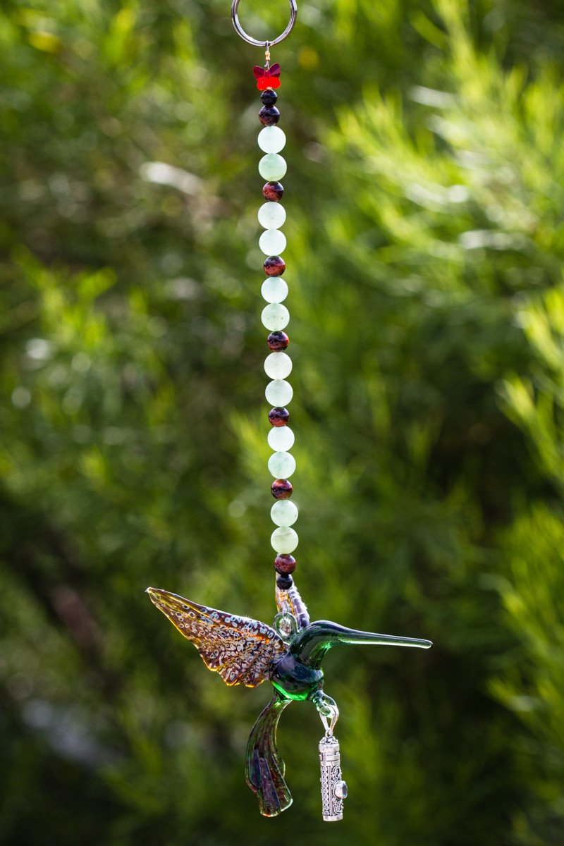 Close up of green glass hummingbird with silver keepsake urn pendant and pale green bead string against a lush garden background.