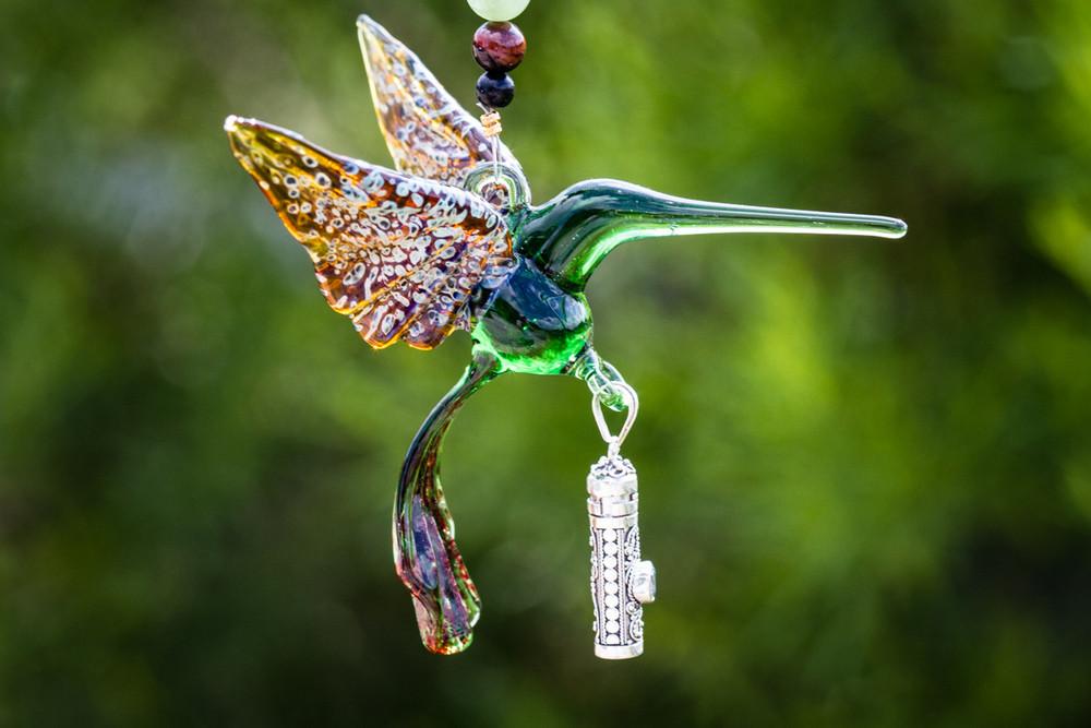 Green glass hummingbird figurine with a silver keepsake urn pendant and a crystal butterfly against a blurred green background