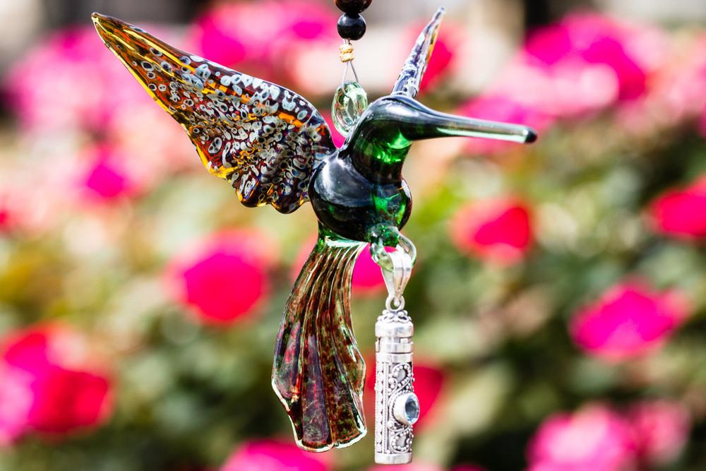 Green and tan glass hummingbird with a silver keepsake urn pendant and beaded accents in front of pink garden flowers